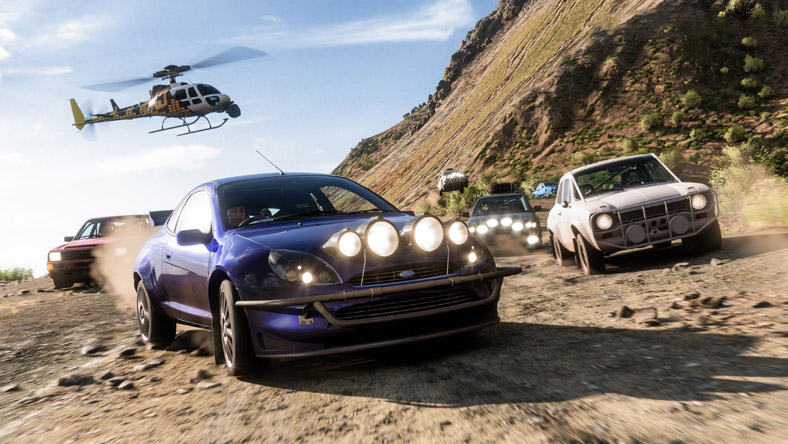 Forza Horizon 5. Um Ford Puma a percorrer uma estrada de terra batida enquanto é perseguido por diversos carros e um helicóptero.