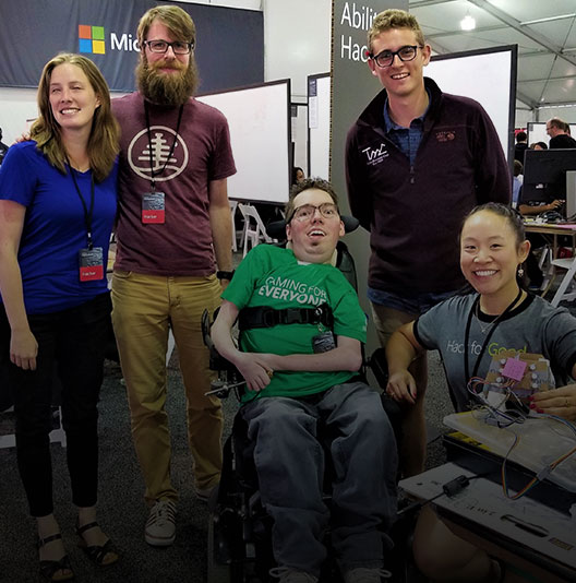 Na tenda Hackathon, uma equipe de cinco pesquisadores, designers e engenheiros posa com um protótipo, todos sorrindo.
