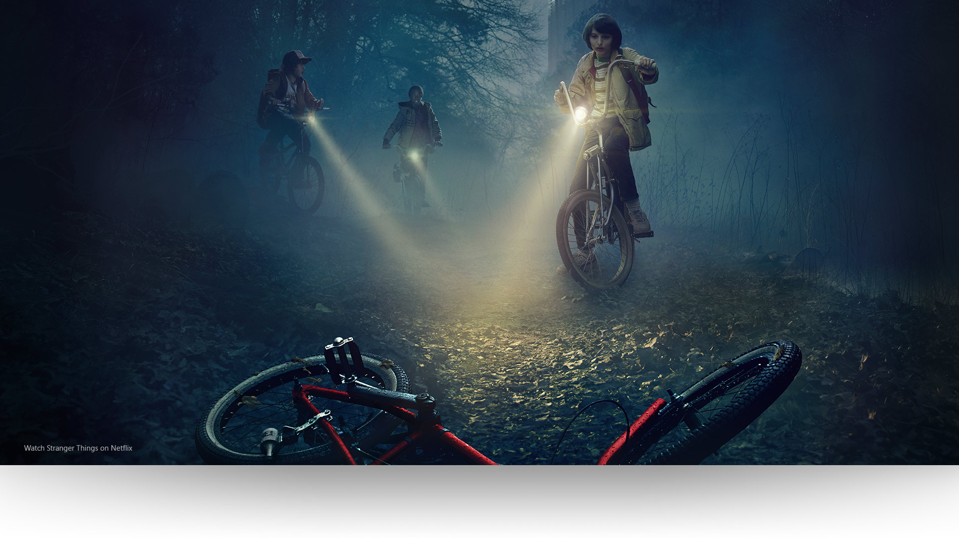 Stranger Things – Dustin, Lucas and Mike shine their lights on an abandoned bicycle on a gloomy forest path. Watch Stranger Things on Netflix.