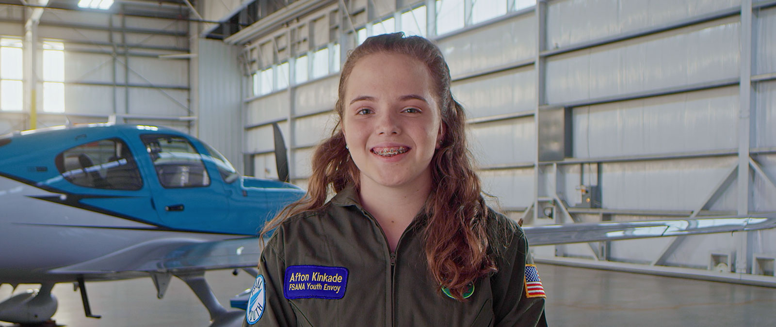 Pilot Afton smiles in a flight suit with a airplane hanger in the background. 