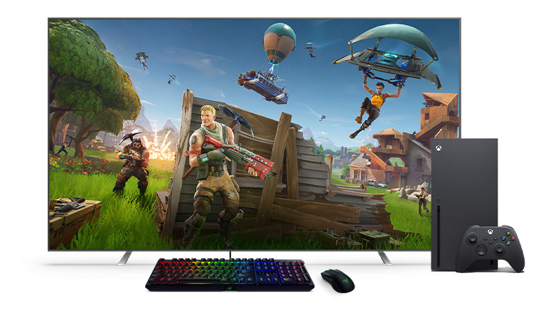 Um teclado preto e um mouse em frente a um Xbox Series X e uma TV.