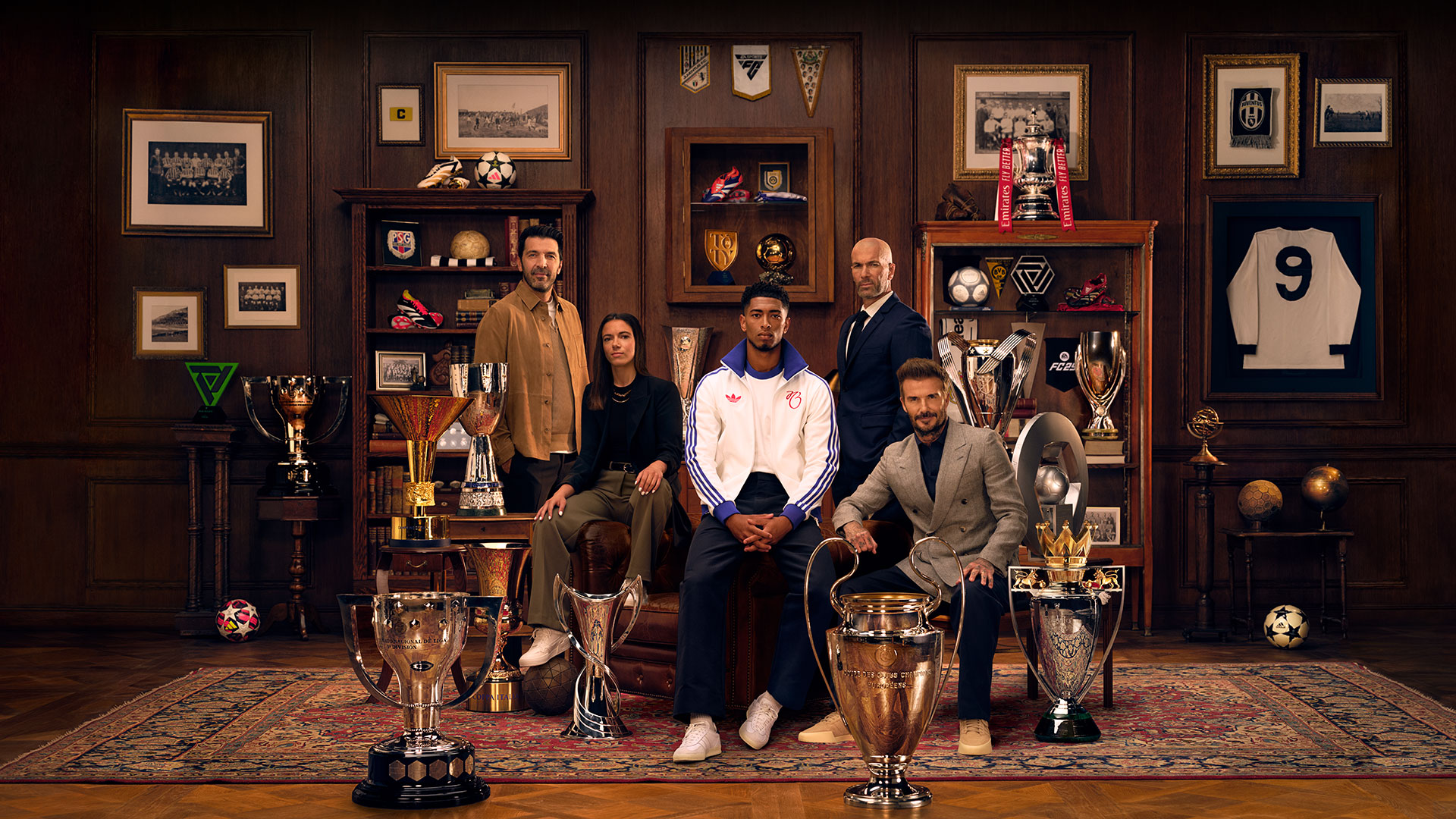 Five people sitting in a club room surrounded by multiple trophies and football memorabilia.