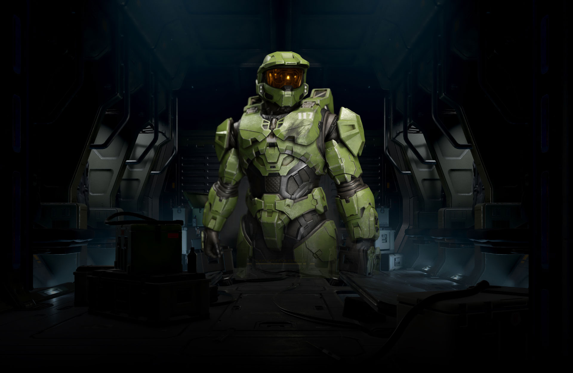Halo Infinite. Master Chief stands in a dimly lit room, the front of his armor slightly damaged under the “117” on his chest.