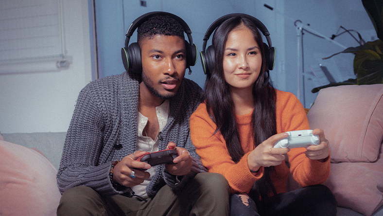 A woman and a man wearing headsets and playing a game with Xbox controllers.