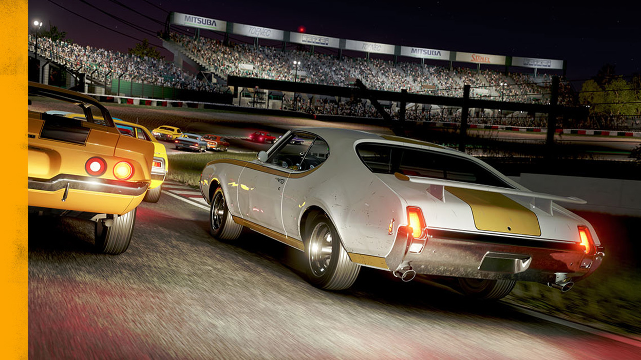 Old muscle cars race around a track at night.