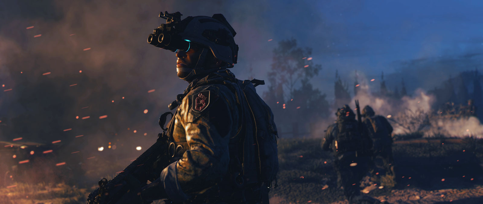 A soldier wearing night vision goggles looks over his shoulder towards distant fire and smoke.