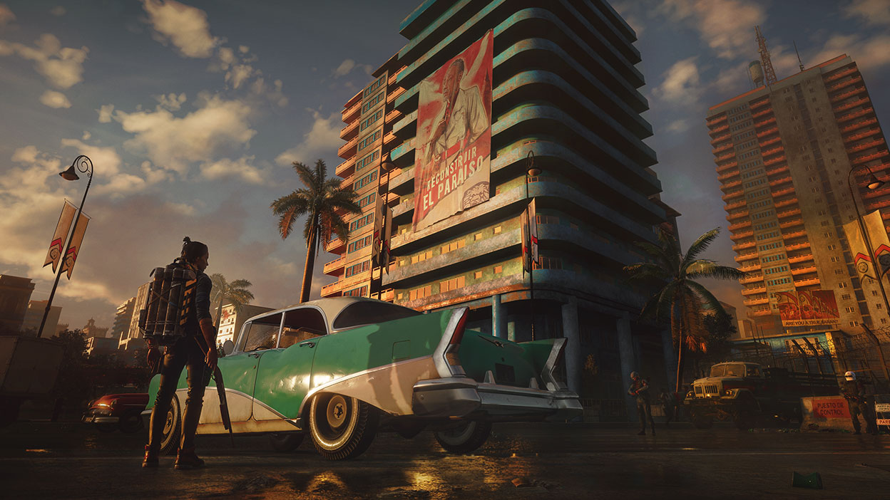 Мужчина с оружием стоит рядом с винтажным автомобилем с возвышающимися строениями на заднем плане в игре Far Cry 6.